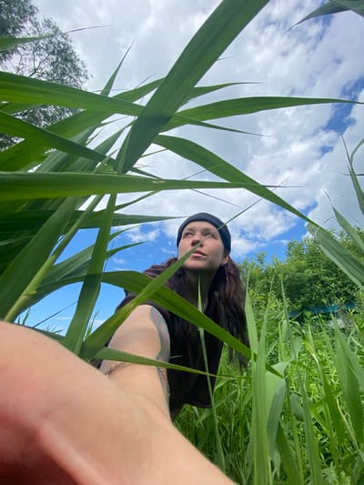 In Petrovskoye, Russia, a low-angle, wide shot captures a person partially obscured by tall green grass. The person, wearing a black beanie and a dark top, looks upwards towards a bright blue sky dotted with white clouds. Their left arm, with a tattoo on the forearm and a silver bracelet, is extended into the foreground, nearly touching the camera lens. The background reveals dense green foliage and trees. The scene appears to be outdoors, possibly in a natural or park-like setting within the city.