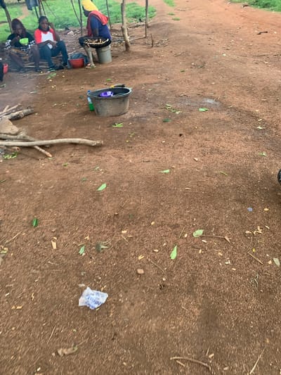 The image depicts an outdoor scene in Buga da Gindi, Nigeria, likely a rural or semi-rural environment given the dirt ground and basic structures. Several individuals are gathered, with some seated on the ground and others partially visible. In the foreground, two women are seated, one wearing a patterned dress and the other a red and white top. To their left, another person is seated wearing a black and white patterned outfit. Further back, a person in a blue garment with red and yellow accents is seated on a bucket, and behind them, another bucket is visible. A large, dark gray bucket with a purple object inside is placed to the right of the seated individuals. There's also a red bucket visible. The ground is predominantly brown dirt with scattered leaves and debris. The lighting suggests it might be daytime, possibly overcast or late afternoon, as there are no strong shadows. The overall atmosphere appears casual and communal, with people gathered together, possibly for a meal or social interaction. A small, crumpled plastic bag with some text is visible on the ground in the middle right of the image.