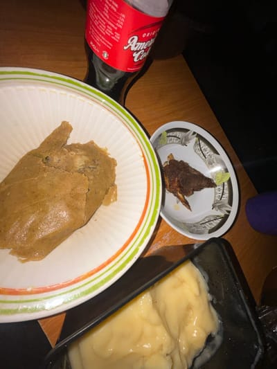 The image depicts an indoor meal setup, reportedly located in Debasco, Nigeria. No individuals are visible within the frame. The primary subjects are various food and beverage items arranged on a light brown wooden surface.
In the upper central portion, a clear plastic bottle containing a dark liquid, identified by its red label with white text as "America Cola" and "25 CL" volume, stands upright. To the left, a large, irregularly shaped mass of light brown, cooked food, consistent with a steamed pudding or paste (e.g., *moin-moin*), is presented in a white ceramic bowl with distinct green and orange stripe patterns along its rim. To the right of this bowl, a smaller, dark brown piece of cooked meat, possibly bone-in and appearing charred or fried, rests within a shallow white ceramic bowl adorned with a black and white geometric pattern and green leaf motifs. In the lower right foreground, a black rectangular plastic container holds a viscous, pale yellow food substance, resembling a starch-based swallow (e.g., *fufu* or *amala*) or a creamy pudding, with some liquid residue visible along its interior edges. The scene is illuminated, suggesting an ambient light source from the left or above.