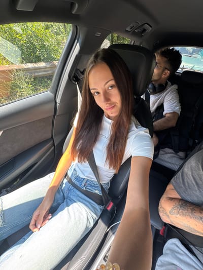The media file shows three people inside a car, likely traveling on a sunny day. The image is oriented vertically, with the camera positioned low in the car. In the foreground, a woman with long, brown hair is looking towards the camera, smiling. She is wearing a white t-shirt and blue jeans, and her seatbelt is fastened.
Behind her, a man with curly dark hair and glasses is visible, appearing to be asleep or resting. He is wearing a white t-shirt and has headphones around his neck. Further back, another person's arm is visible, showing a tattoo on their forearm. They are wearing a gray t-shirt and a seatbelt.
The interior of the car is visible, including black leather seats and a car door. Through the windows, lush green trees and vegetation suggest a rural or natural setting. The bright sunlight filtering in indicates it is daytime. The overall mood appears relaxed and casual, consistent with a road trip. Text is visible on a small rectangular label on the car's interior roof trim, reading "AIRBAG".