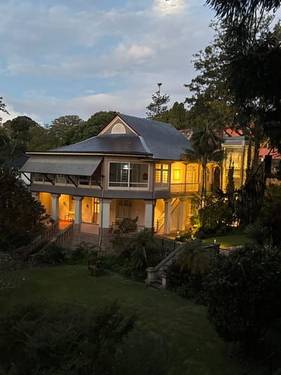 The image depicts a stately, multi-story house at dusk, bathed in warm artificial light emanating from within its windows and from exterior lamps illuminating the grounds. The house, constructed with stone and rendered walls, features a prominent roofline with a gabled dormer window. A covered veranda with support columns runs along the front, leading to a balcony accessible by stairs. Lush green trees and dense foliage surround the property, creating a sense of seclusion. The sky above is a pale blue with scattered, wispy clouds, and a faint full moon is visible in the upper left quadrant. A well-maintained lawn occupies the foreground, with stone steps and pathways winding through the garden. A small, dark-colored statue or sculpture stands on a wooden pedestal in the lawn. The overall impression is one of tranquility and established grandeur within a suburban or semi-rural setting. The location is specified as Woolloomooloo, Australia.