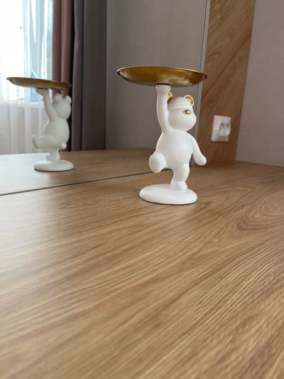 A white ceramic bear figurine holding a gold-colored dish is positioned on a wooden table, reflecting in a mirror behind it. The figurine is facing to the right, with its right arm raised, holding the dish above its head. Its left arm is bent at the elbow, and its left leg is extended backward. The figurine has gold-colored ears and eyes. In the background, a window with sheer white curtains and brown drapes is visible. To the right of the figurine, a white electrical outlet with two circular openings is mounted on a wood-paneled wall. The overall scene is indoors, on a flat surface that appears to be a table or countertop with a wood grain pattern. There is no indication of the city of Meleuz, Russia, in this image.