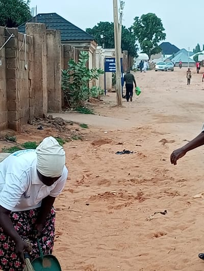 In Wuro Dole, Nigeria, an individual is sweeping sand on a dirt road in front of a fenced property. The person is wearing a white polo shirt, a beige head covering, and a black and floral-patterned skirt. They are using a green dustpan and a broom to clear the sandy ground. In the background, other individuals are visible walking on the road, and several buildings, including a structure labeled "GRAND HOTEL," are present. The environment appears to be an outdoor, semi-urban setting with trees and a cloudy sky.