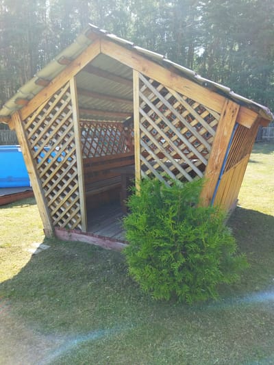 This image shows a wooden gazebo with a metal roof on a sunny day. The gazebo has lattice-work sides and contains wooden benches and a table. In front of the gazebo is a green bush. To the left of the gazebo, a bright blue inflatable swimming pool is partially visible. The scene is set in a grassy yard with trees in the background, suggesting a rural or suburban location in Baburino, Russia. The lighting indicates it is daytime, likely late morning or early afternoon, with the sun casting shadows. There are no people visible in the image, and the overall mood is peaceful and relaxed, suggesting a recreational outdoor setting. There is no visible text in the image.