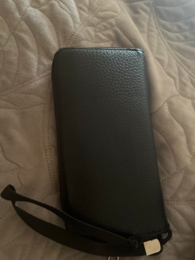 A black, textured wallet lies on a soft, beige blanket. The wallet is rectangular and appears to be made of leather. A black strap is attached to the right side of the wallet, with a small, silver buckle visible at the top of the strap. The blanket has a quilted pattern with wavy lines. The scene is brightly lit, suggesting it might be daytime indoors. There is no visible text in the image.