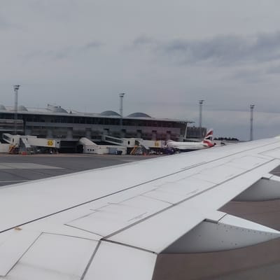 The image captures a view from an aircraft window at Stockholm-Arlanda Airport, Sweden, looking towards a terminal building under an overcast sky. The white wing of the observer's aircraft, featuring visible panel lines and control surface mechanisms, dominates the lower and right foreground. In the midground, a modern airport terminal building is visible, characterized by its multi-level grey facade, large windows, and distinctive curved roof sections. Two jet bridges are prominently featured: one at Gate 65, displaying "65" in yellow on a grey panel, and another at Gate 66, marked "66" in yellow. A British Airways aircraft, identifiable by its white fuselage and red, white, and blue tailfin (Union Flag livery), is parked at Gate 66, partially obscured by the observer's wing. In the background, several tall, slender metal lattice towers, equipped with lights or sensors, rise above the terminal building. The tarmac below is grey, with visible white ground markings. No individuals are discernibly visible within the frame.
