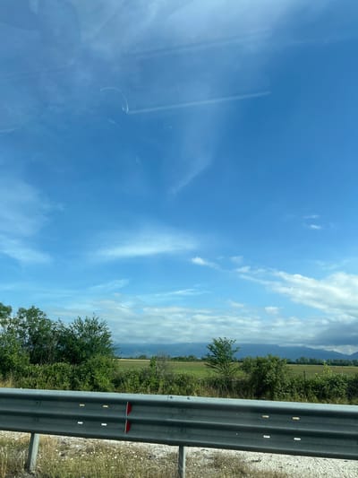 The image captures a bright, sunny daytime scene, likely viewed from a moving vehicle in Malo Konare, Bulgaria. The upper left portion of the frame shows a clear blue sky with some wispy white clouds and light reflections, possibly from a car window. Moving across the sky to the right, more prominent cumulus clouds are scattered, particularly towards the horizon.
Below the sky, the landscape features extensive green fields, indicative of agricultural land, dotted with various trees and bushes. In the distance, a range of mountains or hills with a hazy blue hue forms the backdrop, partially obscured by clouds.
In the immediate foreground, occupying the right side of the image, is a silver metal guardrail, commonly found alongside roads or highways. It runs horizontally across the frame, showing its corrugated structure and several mounting bolts. Two red reflectors are visible on the guardrail, and additional white reflective markings are present on its surface. The ground adjacent to the guardrail appears to be gravelly or dirt. The overall impression is one of a scenic journey through a rural landscape. No visible text is present.