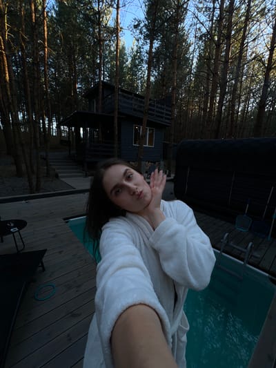 A young woman, wearing a white bathrobe, poses for a selfie in an outdoor setting. She has her left hand near her cheek and her lips are pursed. The background shows a wooden deck, a small swimming pool, a black structure that resembles a sauna, and a modern, dark-colored house or cabin nestled among tall trees. The environment suggests a secluded, wooded location. The image was captured in Ivachiv Horishnii, Ukraine.