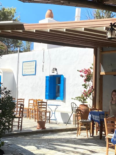 The image depicts an outdoor patio of a restaurant, identified as "ΕΣΤΙΑΤΟΡΙΟ 'ΧΡΥΣΟΠΗΓΗ' ΣΙΦΝΕΪΚΗ ΚΟΥΖΙΝΑ" (Restaurant 'Chrysopigi' Sifnian Cuisine), located in Φάρος, Greece.
The scene is illuminated by direct sunlight, casting distinct shadows across the stone-paved ground. A white-washed building occupies the background, featuring a prominent blue-painted wooden window with open shutters. Above this window, a wall-mounted black lantern is visible. To the left of the window, a rectangular blue sign with white Greek text is affixed to the wall. A white vertical pipe runs along the left side of the building. On the rooftop, a terracotta amphora-shaped pot stands against a clear blue sky.
The foreground and mid-ground are furnished with outdoor dining elements. Numerous wooden chairs, some with slatted backs, are arranged on the patterned stone pavers. Some chairs are grouped, and one small terracotta pot with a green plant is present on the ground. A small, round, light blue metal table with a cross-base is positioned in the mid-ground. To the right, a table is covered with a blue and white patterned tablecloth, surrounded by wooden chairs, one of which has a cushion. A large terracotta pot containing vibrant pink bougainvillea is situated against the building wall on the right.
An overhead pergola structure, composed of multiple dark brown wooden beams, provides partial shade across the top of the image. A hanging lantern with a metal frame and glass panels is suspended from the pergola on the right.
A single female subject is partially visible on the far right side of the image, standing in a brightly lit area. She is facing slightly left, looking towards the mid-ground. Her hair appears light-colored and damp or disheveled. She is wearing a light, possibly sleeveless, top. No discernible action or interaction is depicted beyond her standing presence. Green foliage is visible in the far left foreground and background.