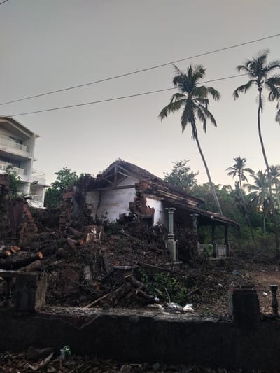 The image depicts a partially destroyed building in what appears to be a state of disrepair. The structure is surrounded by debris and overgrown vegetation. Architectural details, such as pillars and roof framing, are visible despite the damage. In the background, palm trees stand tall against a pale sky. A modern building is partially visible in the lower part of the frame. Power lines stretch across the upper left portion of the image. The specific location in Marna, India, cannot be determined from the visual information alone.