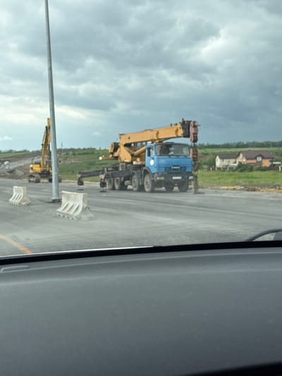 The image shows a scene in Shchepkin, Russia. A blue and black crane truck with a yellow crane arm is parked on the right side of a road. Adjacent to the crane are white and gray construction barriers. Further down the road, a yellow excavator is visible. The background includes a grassy field and houses. The image appears to be taken from inside a vehicle, with part of the vehicle's interior visible on the right side. The sky is cloudy.