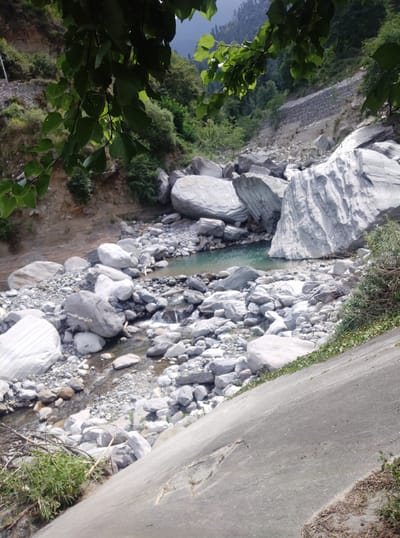 This image captures a rocky riverbed in Baragran, India. In the foreground, a large, light gray, smooth rock slopes upwards from the bottom right, with dry grasses and some green vegetation along its edge. The middle ground is dominated by a shallow, turquoise body of water, bordered by numerous large, smooth, gray boulders of various sizes. The river flows from the left towards the center of the image, with smaller rocks and pebbles lining its banks. In the background, a steep, rocky hillside rises, with dense green trees and vegetation covering parts of it. A stone retaining wall is visible partway up the hillside, suggesting human intervention in stabilizing the slope. Overhanging the scene from the top are dark green leaves and branches, obscuring a portion of the sky and upper hillside. The overall impression is one of a natural, rugged landscape with evidence of human presence.