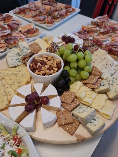This is a high-angle, medium shot photograph of a cheese board at what appears to be a catered event. The cheese board is round and made of light-colored wood, filled with various cheeses, crackers, grapes, olives, and nuts. There are several types of cheese, including wedges of brie, sliced cheeses with herbs and spices, and blue cheese. Crackers are scattered amongst the cheeses, some with seeds. A small white bowl contains a mix of almonds and cashews. Green grapes and black olives are also present. In the background, trays of small appetizers or bruschetta are visible on white platters. A white bowl containing a salad is partially visible in the lower left corner. The overall setting suggests a gathering or party, possibly in a venue or a home in Garbsen, Germany. There are no people clearly visible in the image.