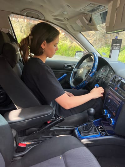 An indoor shot captures a young woman with dark hair in a ponytail, wearing a black t-shirt, seated in the driver's seat of a car. She is looking down at the car's center console, with her right hand near the gear shift. The car's interior features blue decorative accents on the steering wheel and dashboard. Outside the car, a rocky, overgrown hillside is visible through the windows, bathed in sunlight. This image was taken in Pechinska, Bulgaria.
