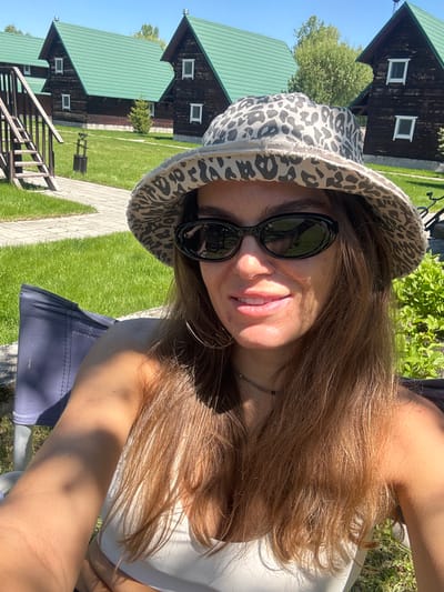 A young woman with long, light brown hair and sunglasses poses for a selfie. She is wearing a white top and a spotted bucket hat. In the background are several identical wooden cabins with green roofs, set amidst a grassy lawn and paved walkways. This scene is in Aya, Russia, as indicated by the location metadata.