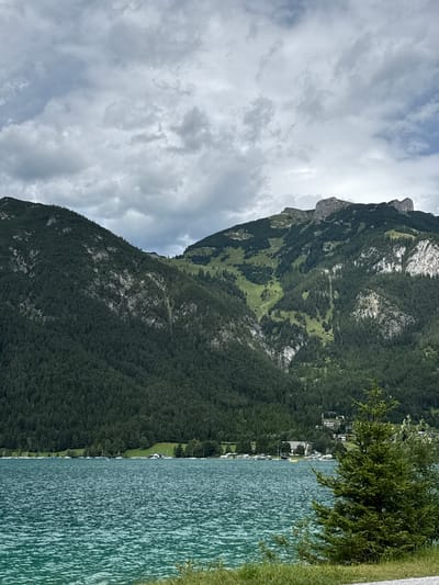This is a photograph of the Achensee lake, with mountains in the background, taken from the shore in Pertisau, Austria. The foreground is dominated by the turquoise waters of the lake, which show ripples and waves. A few green trees and grass are visible along the bottom edge of the frame. In the middle ground, a line of sailboats and smaller vessels are moored along the shore, with buildings and trees forming a backdrop. The background consists of large, densely forested mountains, with a lighter green patch of grass or a ski slope visible on one of the slopes. The peak of one mountain features a distinctive rocky formation. The sky is overcast with dramatic, grey clouds.