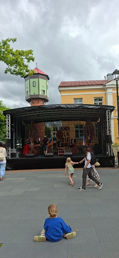 A daytime outdoor scene captures a folk music festival in Viljandi, Estonia. A band performs on a raised stage adorned with a banner reading "VILJANDI FOLK MUSIC FESTIVAL." The stage is backed by a building with yellow siding and a red roof. To the left of the stage stands a distinct green and brown tower with a red roof, characteristic of Viljandi's historical architecture. In the foreground, a young child with blonde hair sits on the paved ground, facing away from the camera, wearing a blue sweatshirt and light-colored shoes. To the right, a barefoot young girl in a green dress holds hands with a person wearing a white t-shirt, dark pants, and a backpack, as they walk across the paved area. Further to the left, a person in light blue pants and a white top with a backpack walks away from the camera. The sky is overcast.