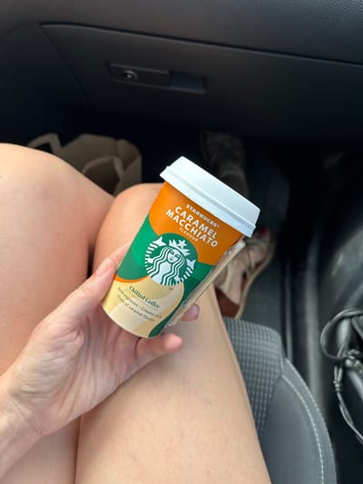A person's hand holds a Starbucks Caramel Macchiato chilled coffee drink in a car. The drink is in an orange and green cup with the Starbucks logo. Text on the cup reads "STARBUCKS® CARAMEL MACCHIATO FLAVOUR" and "Chilled Coffee Bold espresso Creamy milk Hints of caramel flavour". The person's legs are visible, wearing a light-colored skirt or shorts. In the background, a car's interior is visible, including a dark grey seat with stitching and a glimpse of a person's shoes. The scene suggests a casual outing or travel. The lighting indicates it is daytime, possibly warm weather given the attire. The location context is Očinići, Montenegro.