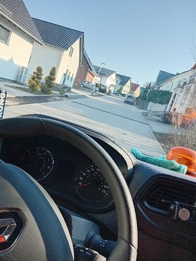 This image depicts a residential street in Alzey, Germany, viewed from the driver's perspective within a stationary vehicle. In the immediate foreground, the vehicle's interior features a black steering wheel, partially obscuring the instrument cluster which shows a tachometer and speedometer at rest. A control stalk extends from the steering column. On the dark dashboard, a teal utility cloth and an orange rotating beacon light are present, alongside a black magnetic phone mount attached to an air vent. A portion of a cup is visible on the far left.
The scene outside the vehicle is a paved residential street with sidewalks, lined on both sides by two-story detached houses. These houses feature dark, pitched tile roofs and predominantly light-colored facades, some accented with brickwork. Landscaped front yards with shrubs, including topiary, are visible. Several parked vehicles, including a silver hatchback and a dark sedan, are positioned along the street. Streetlights are also present. The environment is characterized by a clear, bright blue sky and strong daylight, which creates well-defined shadows. No individuals are clearly discernible within the scene.