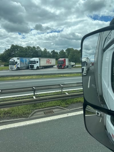 The image shows a view from a vehicle on a highway, likely in or near Weischlitz, Germany, given the context provided. The foreground features the road surface and a guardrail. In the mid-ground, several large trucks are visible, driving in the same direction. One truck has "ARC LOGISTICS" written on its side. In the background, there are trees and a cloudy sky. The scene appears to be a traffic situation on a multi-lane highway. The reflection in the side mirror of the vehicle shows a white truck, also on the highway.
