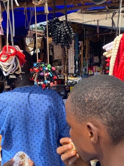 In Nguzu Edda, Nigeria, two individuals are at a market stall adorned with various beaded items. One person, seen from the back wearing a blue shirt with black polka dots, is partially obscured by their own shoulder and torso. In front of them, a second individual, a young male, is shown in profile from the right, facing left and eating. The market stall itself is set up under a makeshift roof, with a blue tarp visible in the background. Hanging from the stall are numerous strands of beads, including black, red, white, and multi-colored varieties. Some strands are clustered together, forming spherical shapes, while others hang in long strings. One cluster of black beads has a tag with the word "JESUS" printed on it. The overall scene depicts a bustling market environment with an emphasis on the sale of beaded accessories.