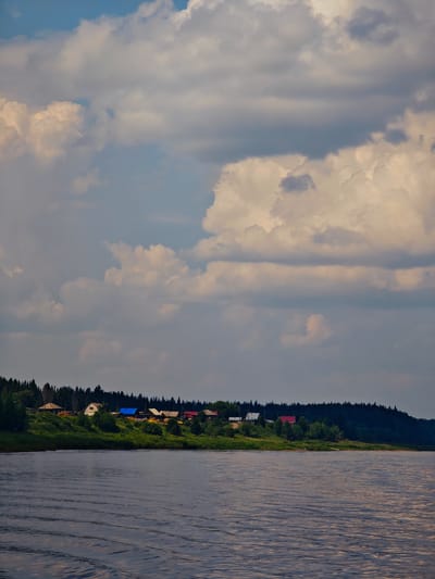 The image depicts a wide, dark river in the foreground, with visible ripples on its surface. The middle ground shows a lush green riverbank with a line of trees forming a dense forest in the background. On the riverbank, nestled among the greenery, is a small village. Several houses with colorful roofs, including red, blue, and white, are visible. The sky above is dramatic, filled with a mixture of bright white and dark gray clouds, suggesting a dynamic weather system. The overall scene is a natural landscape with a touch of human habitation, likely in a rural setting. This scene is consistent with the appearance of the settlement of Ust-Kem, Russia, given its location on a river and surrounded by forests.