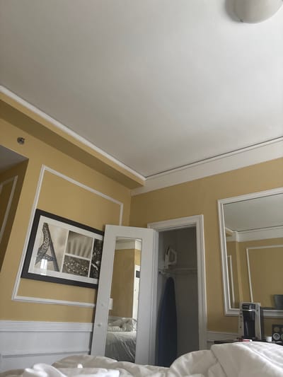 The image depicts a hotel room with yellow walls and white trim. In the foreground, white bedding is visible. To the left, a framed picture of the Eiffel Tower is hanging on the wall. A white door is open, revealing a closet with an ironing board and an iron. To the right, a mirror reflects a coffee maker and a Keurig machine on a counter. The ceiling is white and features a circular light fixture. The scene is indoors, and no people are present. The exact location in San Francisco is not discernible from the image.