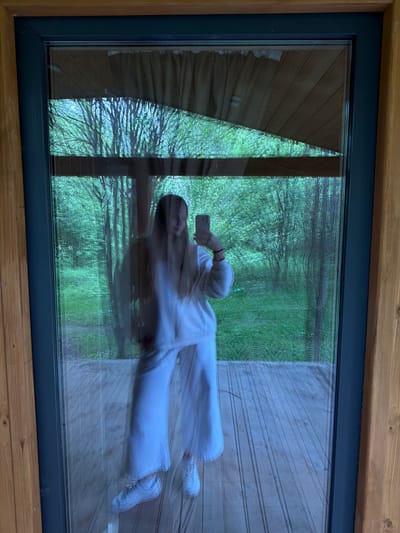 The image captures a scene through and reflected in a large window, framed by light-colored wood. The window itself has a dark grey frame.

Reflected in the window, a person, seemingly female, is visible, standing upright and facing towards the viewer. She has long, light-colored hair and is dressed in a light-colored, possibly white, long-sleeved top, wide-leg trousers, and white sneakers. She holds a smartphone with a light-colored case up in front of her, appearing to be taking a picture or video, or looking at the screen. There is significant motion blur, particularly around her head and upper body, suggesting movement during the exposure.

Beyond the reflection, through the glass, a lush green outdoor environment is visible. This includes numerous trees with dense foliage and green grass, indicating a natural setting, likely during spring or summer. To the right of the reflection, outside the window, a wooden deck or planked floor is visible. To the left, part of an interior space, possibly a curtain or another wall, is reflected, along with some wooden paneling.

The lighting suggests daytime, with diffused light illuminating the scene. No discernible text is visible in the image. The overall impression is of someone capturing a moment in a serene, natural environment from inside a building with large windows.