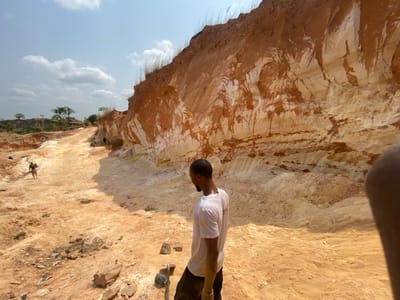 The image portrays an active open-pit excavation site in Asaga Amangwu, Nigeria, under a bright sky with scattered white clouds. A prominent, tall wall of exposed earth dominates the right side of the frame, revealing distinct geological strata of reddish-brown, orange, and light tan material. Sparse dry grass is visible along the top edge of this excavated wall.

In the foreground, a male individual with dark skin and short dark hair is partially visible, facing away from the camera and looking towards the left. He wears a white short-sleeved t-shirt and dark trousers. The ground in front of him is rough and uneven, composed of reddish-brown soil with scattered rocks and a dark-colored tool. A partial view of a human thumb is present in the extreme lower right corner.

Extending into the midground and background is a wide, excavated pathway or trench, consisting of reddish-brown and light tan soil. Along this path, a second male individual is observed walking towards the left, carrying a dark object over his left shoulder. Further in the distance, near the base of the excavated wall on the left, several other indistinct figures are visible, appearing to be engaged in work-related activities. The background features elevated terrain with sparse green trees under the bright sky. The scene is brightly illuminated by direct sunlight.