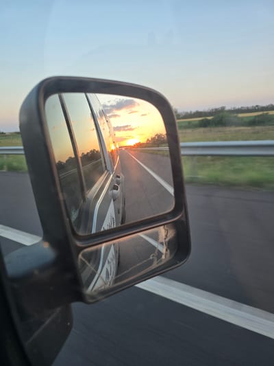 A side-view mirror of a vehicle reflects a sunset over a highway. The mirror frames a road with a white line, and the sky is a pale blue fading to orange and pink at the horizon. The sun is a bright orange orb setting behind a distant tree line. The reflection in the mirror shows the side of the vehicle, with a white decal visible. The road appears to be in a rural area, as fields and trees are visible on either side. The scene suggests a journey on a highway during golden hour. The image is taken from within the vehicle, looking out at the side-view mirror. There is no visible indication of the location being the city of Makó, Hungary.
