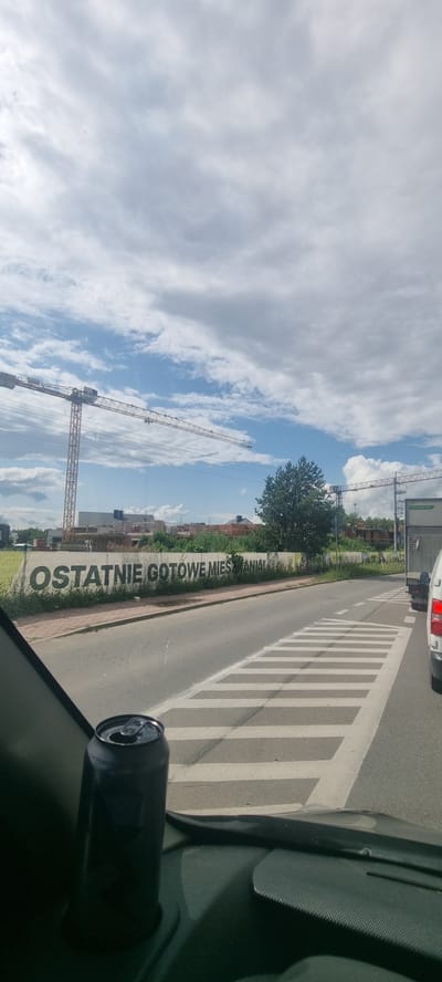 In Otrębusy, Poland, a concrete wall along a road displays large, dark lettering that reads "OSTATNIE GOTOWE MIESZANIA". The wall is situated behind a sidewalk and some vegetation. The scene appears to be a construction area, with cranes visible in the background, suggesting ongoing development. A road with white crosswalk markings is in the foreground, with traffic lanes visible. A dark-colored beverage can rests on the interior dashboard of a vehicle, partially obscuring the view of the road ahead. The sky is partly cloudy, with a mix of blue sky and white clouds.