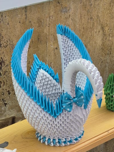 The image shows a 3D origami swan, primarily white with blue accents along the edges of its wings and tail, and a blue bow with white stars around its neck. The swan is placed on a surface composed of wooden planks and a wall with a textured beige surface. A small portion of another origami structure, green in color, is visible in the upper part of the frame. The precise location within Srebarna, Bulgaria, cannot be ascertained from this image.