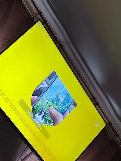 The image shows a close-up, angled view of a bright yellow screen, likely a tablet or monitor, displaying a photograph within its display area. The photograph features a young woman with dark hair and sunglasses, wearing a white t-shirt, standing outdoors with lush green foliage in the background. A hand holding a mobile phone is visible in the foreground of the photograph, capturing the image of the woman.
In the upper left corner of the yellow screen, a small icon resembling a speaker with sound waves is visible, alongside some text in what appears to be Russian.
Along the bottom left edge of the yellow screen, more text in Russian is present, which appears to be a disclaimer or promotional material.
In the bottom right corner of the yellow screen, a small, light-colored square contains the text "OK" with a checkmark.
The overall scene suggests a person is using a digital device to view or share a photograph. The location of Кондоль, Russia, is not depicted in the image itself.
