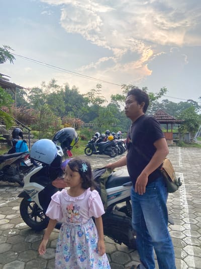 A medium shot, taken at an oblique angle, shows a man and a girl next to several motorcycles in Penanggungan, Indonesia. The man, wearing a black t-shirt and blue jeans, stands over one of the motorcycles, facing the camera. Next to him, a girl in a pink and blue floral dress looks downward. Several other motorcycles are parked in the background. Helmets rest on the seats and handlebars of the motorcycles, reflecting sunlight. The setting is a paved area with a small garden and building visible in the background. The sky shows a mix of clouds and clear areas.