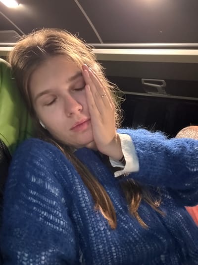 A woman with fair skin and light brown hair is lying down with her hand covering her forehead. She is wearing a textured blue sweater over a white shirt. She is resting on what appears to be a green surface. A window frame with a dark background is visible in the background. There is no specific information in the image to determine the location is Cacak, Serbia.