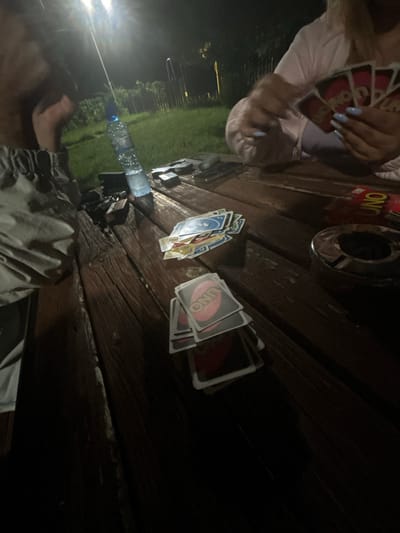 The image shows a group of people playing cards outdoors at night, likely in Dolna Banya, Bulgaria. The scene takes place on a wooden table illuminated by a bright light source overhead. Several UNO cards are scattered across the table, suggesting a game in progress. Among the objects visible are a plastic water bottle, a pack of cigarettes, and an ashtray. One person's hand, adorned with light blue nail polish, is shown holding several UNO cards, indicating active participation in the game. The background reveals a grassy area with some fencing, consistent with an outdoor setting in a suburban or rural environment. The overall atmosphere appears casual and social.