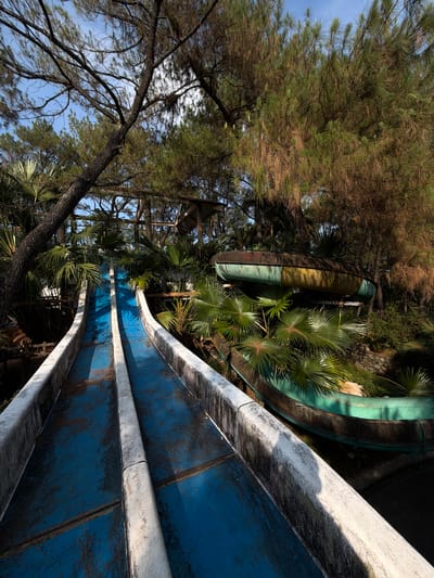 The image depicts an abandoned water park in Thủy Xuân Ward, Vietnam. Dominating the foreground is a blue water slide with white edges, its surface showing signs of weathering and staining. The slide is surrounded by lush, overgrown vegetation, including palm trees and pine trees, partially obscuring the slide's structure. Further in the background, one can see the rusty framework of a water slide tower and other decaying water slides, indicating the extent of the abandonment. The scene is bathed in sunlight, but the overgrowth creates shadows, contributing to the derelict atmosphere.