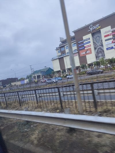 A view from a moving vehicle shows Spur mall and Verona Hotel under a cloudy sky. Various businesses are advertised on the building's exterior. A metal guardrail is prominent in the foreground, obscuring the road and landscape. Cars are parked and moving on the street. Other buildings are also visible in the distance.