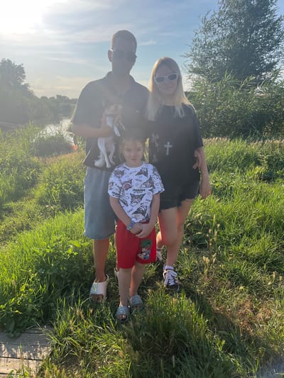 The image captures a family of three and a dog posing for a photograph in a grassy outdoor setting. A man in the center holds a small white and brown dog. He is wearing sunglasses, a dark t-shirt with a graphic print, and light blue denim shorts. To his right stands a young woman with blonde hair, also wearing sunglasses, a dark t-shirt with a white cross graphic, and black denim shorts. In front of the man and slightly to the left is a young girl with dark hair, wearing a white t-shirt with cartoon graphics and red shorts. She has a blue bracelet on her left wrist. All three individuals are looking towards the camera. The background features a body of water on the left, with trees and tall grass. The sky is bright, indicating it is daytime. The overall scene suggests a casual outdoor gathering in a rural or semi-rural environment, possibly near a body of water. The location is identified as Yanovo-Grushevsky, Russia.