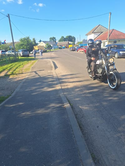 The image captures a street scene in Блеўчыцкі сельскі Савет, Belarus. In the foreground, two individuals, a man and a woman, are riding a motorcycle. Both are wearing helmets. The man is driving the motorcycle, and the woman is seated behind him. They are dressed in casual clothing. The motorcycle is dark in color and appears to be a cruiser style. The road is paved and has a sidewalk on the left side. In the background, there are several houses with red and white roofs, a few parked cars, trees, and a utility pole with wires. The sky is clear and blue, indicating a sunny day. The overall environment suggests a rural or suburban setting within Belarus.