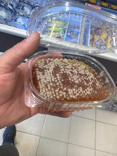A person's hand holds a clear plastic container filled with honeycomb. The honeycomb is a rich amber color, with visible hexagonal cells. The container is open, revealing the honeycomb's textured surface and the honey within. In the background, a refrigerated display case is visible, filled with various products, some of which are wrapped in yellow and blue packaging. The floor appears to be tiled. The scene is indoors, likely in a store or market, given the presence of the display case and the tiled floor. The lighting suggests it is daytime. The image is a close-up of the honeycomb, with the hand and the background slightly out of focus, emphasizing the product. The overall impression is of a food item being presented or examined.