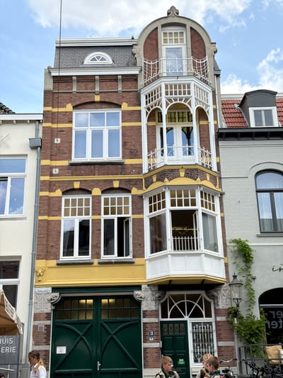 The image shows a multi-story building on a street in Roermond, Netherlands. The building is made of brick with yellow decorative bands and features several white-trimmed windows and a white bay window. The ground floor has large green double doors. To the left of the building, a portion of another building is visible with a yellow awning and signage. On the right side of the building, a lamppost is attached to a lighter-colored building with ivy growing on it. Three young people are visible in the foreground, one standing to the left and two standing near the green doors. Bicycles are parked to the right. The sky is cloudy.