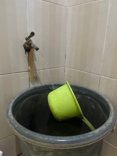 In Anggaswangi, Indonesia, a close-up shot captures a corner of a bathroom with tiled walls. A metal faucet, old and weathered, is dripping water into a large, dark grey plastic bucket that is filled with dark water. A bright green plastic ladle is submerged in the water within the bucket, its handle angled upwards. The wall tiles are light beige with vertical darker stripes, and some water marks are visible on the wall around the faucet and where the bucket meets the wall.
