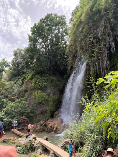 In Anna, Spain, a natural waterfall cascades down a rocky, verdant cliff face. A shallow body of water, possibly a river or pool, flows in the foreground, crossed by a wooden boardwalk. Several individuals are visible: a group of three women and a man stand on the boardwalk near the left side of the frame, looking towards the waterfall. Further along the boardwalk, a man and a woman, both in swimwear, are positioned near the center. Another person, partially visible, stands further down the boardwalk on the right. Dense foliage, including trees and hanging vines, frames the waterfall and surrounds the area. The sky above is overcast with scattered clouds.