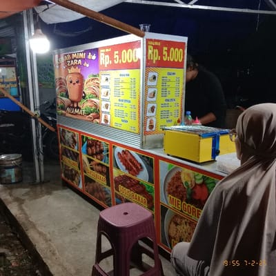 A food stall at night advertises "Kebab Mini Zara" and various grilled meats like "Sosis Bakar" and "Bakso Bakar" along with "Mie Goreng". Prices are listed as Rp. 5,000. A woman wearing a headscarf stands near the stall, where a vendor appears to be preparing food. A red plastic stool is positioned beside the stall. The timestamp "19:55 7-2-26" is visible in the upper right corner.