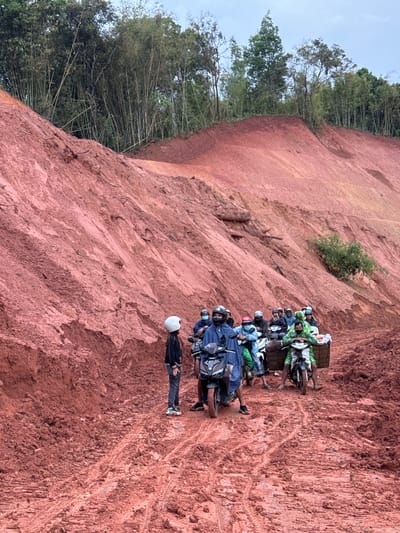 A group of people on motorcycles are gathered on a muddy red dirt road in Nao Sẻ, Vietnam. The road is surrounded by steep, reddish-brown dirt hills, likely the result of recent excavation or erosion. Lush green trees and bamboo grow on the higher slopes and in the background. Several individuals are seated on motorcycles, some wearing helmets, while others stand beside their vehicles. The mud on the road is deeply rutted, indicating recent vehicle traffic. The sky appears overcast.