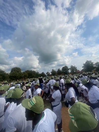 The image depicts a large gathering of young adults, predominantly wearing white t-shirts and green caps with white and green stripes. They are assembled outdoors on a dirt ground, with a line of trees and some low-rise buildings visible in the background. The sky above is partially cloudy. The scene suggests an organized event or activity involving a significant number of participants, likely in a rural or semi-urban setting. The presence of matching uniforms implies a specific group or program. The location is identified as Ise- Ekiti, Nigeria.