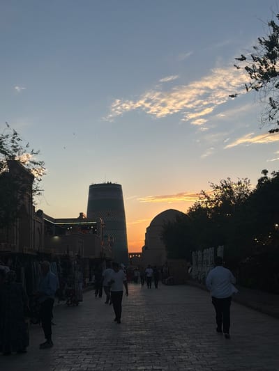 In Khiva, Uzbekistan, an evening street scene unfolds at dusk, with the Kalta Minor Minaret and a domed structure silhouetted against a pastel sky. The minaret, a distinctive cylindrical tower with intricate mosaic patterns, stands prominently, while a smaller dome, likely part of a madrasa, is visible to its right. Scattered clouds, tinged with the warm hues of the setting sun, drift across the sky.

The foreground of the image is a cobblestone pathway, lined with market stalls on the left and a dense foliage of trees on the right. The stalls display an assortment of goods, likely textiles or souvenirs, and a few individuals are browsing or tending to the merchandise. People walk along the path, their figures cast in silhouette by the fading light. One man in a light-colored shirt and dark pants walks away from the viewer, his stride suggesting he is in motion. Another individual in a white shirt and dark pants walks towards the viewer. The overall atmosphere is one of a bustling marketplace winding down for the day, with a sense of tranquility as the day transitions into night.