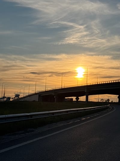 The image captures a sunset over a highway in Sukkolovo, Russia. The sun, a brilliant orb of light, is low on the horizon, casting a warm, golden glow across the sky and clouds. Wispy cirrus clouds are streaked across the sky, illuminated by the setting sun. A highway overpass is visible on the left, with streetlights lining its edge. The road surface on the right is dark and reflects the dim light. The scene conveys a sense of tranquility and the beauty of nature as day turns into night. The time of day is sunset, and the weather appears clear with some cloud cover.