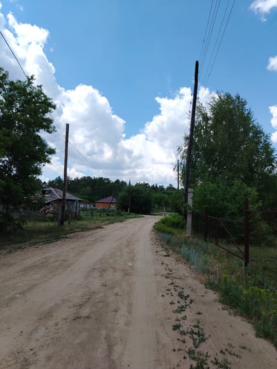 A dirt road curves through a rural landscape under a bright blue sky dotted with fluffy white clouds. To the left of the road, a wooden fence encloses a yard with a pile of logs and a rustic wooden house. Further back, another house with a reddish-brown roof is visible. On the right side of the road, a rusty metal fence runs along the perimeter, bordered by overgrown grass and weeds. Several wooden utility poles with wires are positioned along the road, disappearing into the dense green trees that line the horizon. The overall scene depicts a quiet, undeveloped area, possibly in a village, with a clear, sunny day.